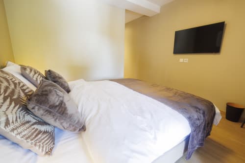 Chambre moderne à La Couronne Boutique Hôtel à Mies avec lit blanc, oreillers gris, murs jaunes, TV murale