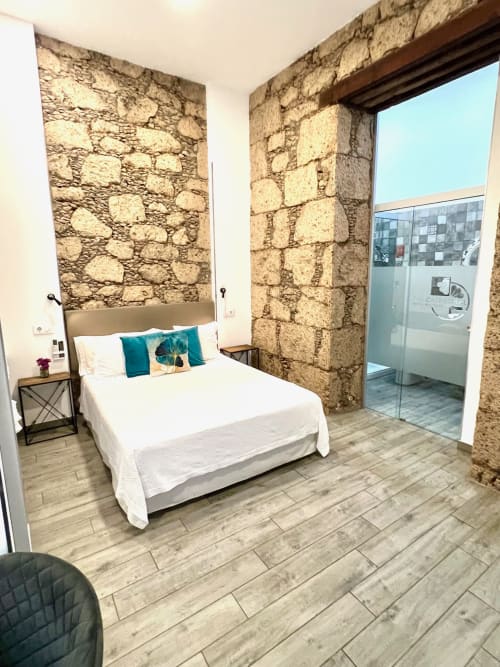 Modern double bedroom with exposed stone walls, white bed with teal pillows, wooden flooring, and ensuite bathroom at Hotel