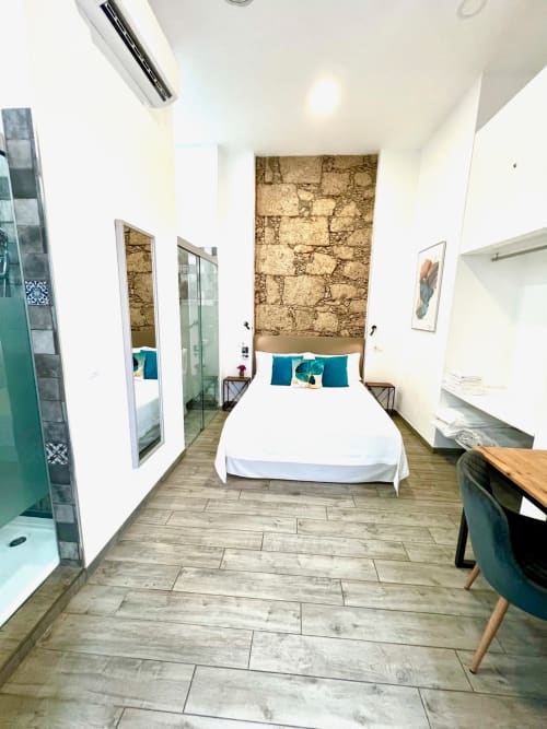 Habitación moderna en The Cathedral Vegueta con cama doble, almohadas azul verdoso, pared de piedra, suelos de madera y baño privado