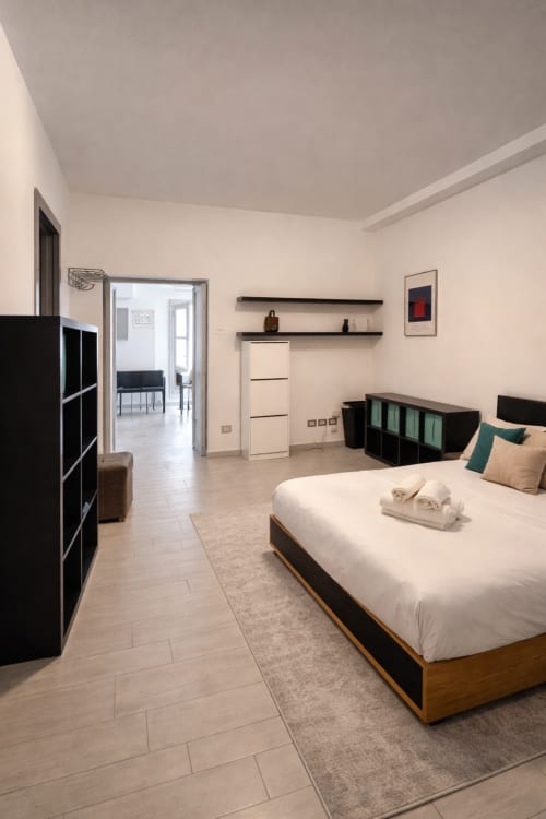 Modern Milan apartment bedroom with queen bed, black storage, shelving, and open-plan living area with city views