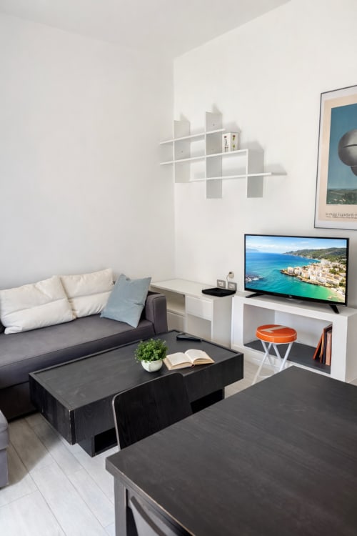 Modern Milano apartment living room with sofa, flat-screen TV showing coastal view, black coffee table, and white wall