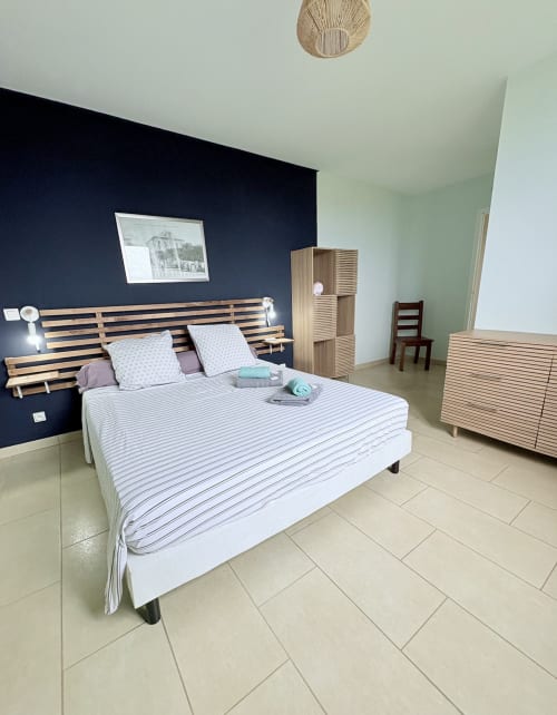 Chambre moderne à La Conciergerie Martinique avec lit à lattes bois, linge rayé, mur d'accent bleu marine et décor minimaliste