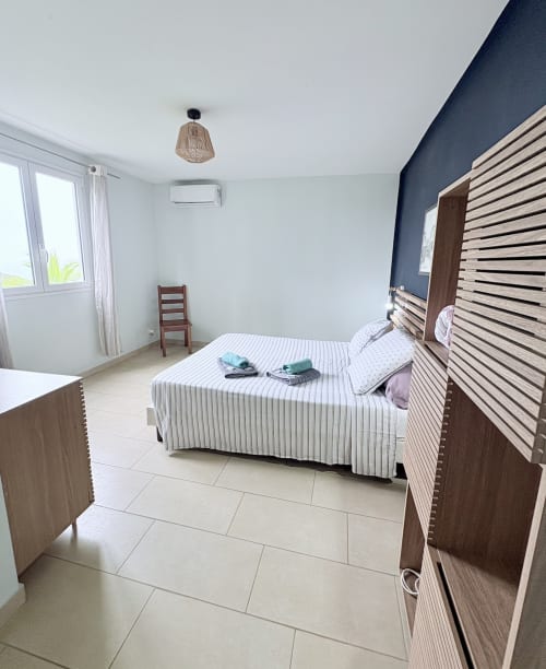 Chambre moderne à La Conciergerie Martinique avec lit rayé, mur d'accent sombre, mobilier en bois et balcon vue mer