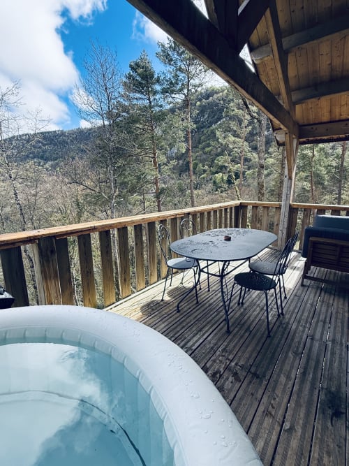 Terrazza in legno con vasca idromassaggio, tavolo da pranzo e vista sulle montagne boschive del lodge Cabanes et lodges du Belvédère