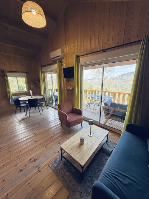 Interno di lodge moderno con soffitti in legno, vista lago attraverso porte vetrate, tavolo da pranzo e zona soggiorno presso Cabanes et