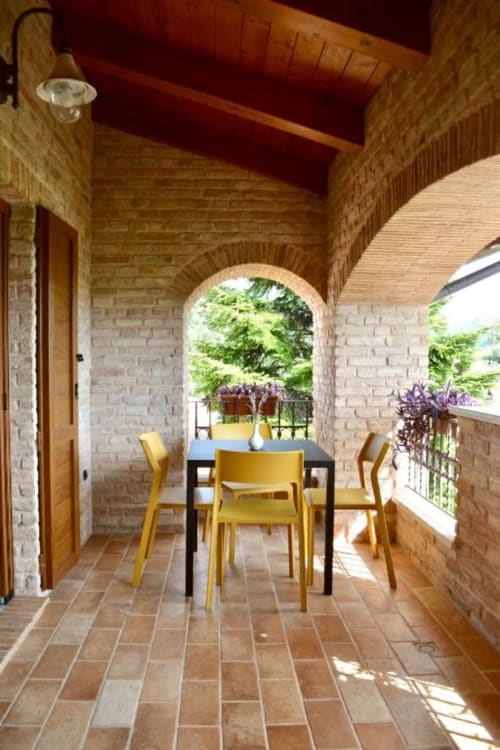 Brick covered porch with yellow chairs at dining table overlooking garden at Agriturismo Colle Mura, Gualdo Tadino
