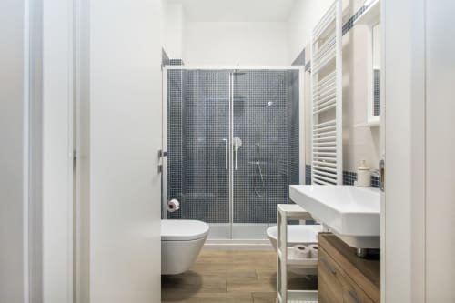 Современная ванная комната в Magnolia Apartments Milan с темной плиточной душевой кабиной, белой раковиной, унитазом, электрическим полотенцесушителем и деревянным полом