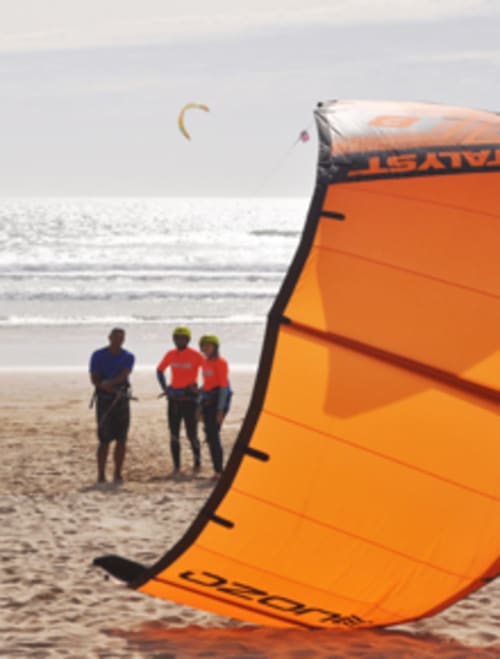 Aula de kitesurf na praia da Costa da Caparica com pipa laranja e três instrutores em fatos de neopreno
