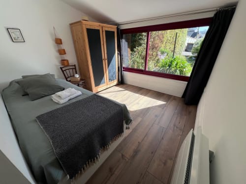 Chambre lumineuse à Destination Étretat avec vue sur le jardin, lit en bois, armoire et grandes fenêtres donnant sur la verdure
