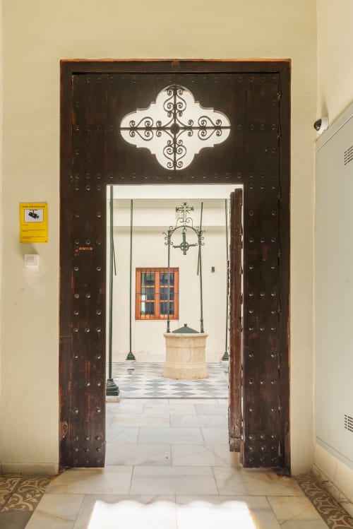 Puertas de madera oscura ornamentadas abriéndose a un patio con cruz de hierro forjado y fuente, apartamento Jerez Centro, Puerto Santa