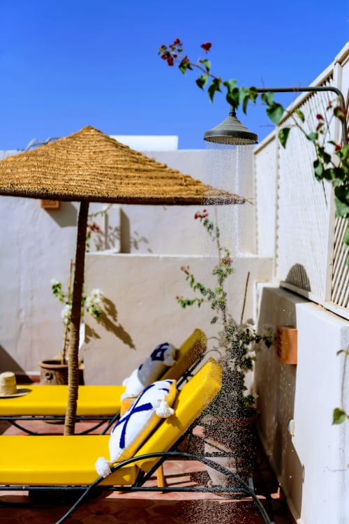 Douche du patio du riad avec chaise longue jaune, parasol en paille et plantes grimpantes sur murs blancs
