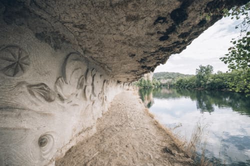 Surplomb de falaise calcaire avec détails sculptés dominant une rive sablonneuse et une rivière calme, Saint-Cirq-Lapopie, France