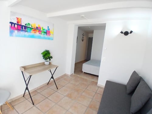Modern hotel room at Hôtel Quai d'Azur with colorful cityscape art, wooden desk, green plant, gray sofa, and terracotta tile