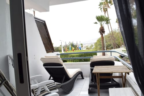 Modern apartment terrace in Costa Adeje with lounge chairs, wooden table, palm trees, and city views overlooking pool area