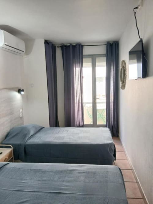 Twin beds in bright hotel room with dark curtains and balcony doors at Hôtel Quai d'Azur, Le Grau-du-Roi