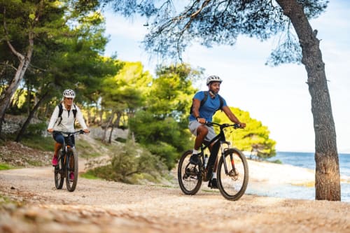 Dois ciclistas pedalando em uma trilha costeira perto da Vila 5 em Vila do Conde, Portugal, com praia e pinheiros.