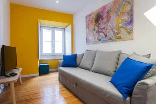 Apartamento luminoso no Bairro Alto com parede de destaque amarela, sofá cinzento com almofadas azuis, piso em madeira e arte colorida.