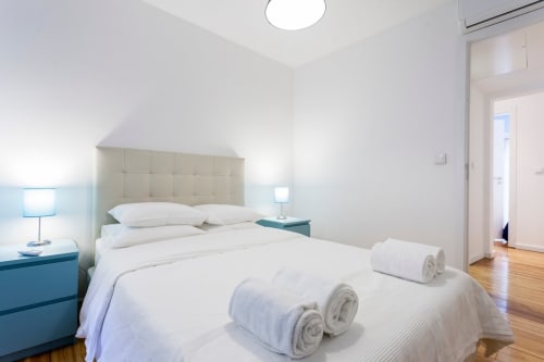 Quarto moderno no Bairro Alto com cama de casal, mesas-de-cabeceira teal, roupa de cama branca, toalhas enroladas e piso em madeira