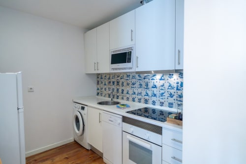 Cozinha branca moderna em apartamento lisboeta com painel de azulejos azuis, micro-ondas, fogão de indução e máquina de lavar.