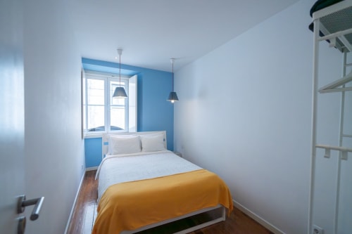 Quarto moderno e luminoso com parede azul, manta amarela e lustres suspensos no apartamento do Bairro Alto, Lisboa.