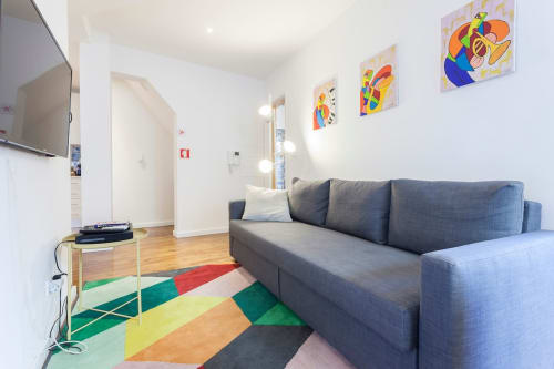 Sala de estar moderna com sofá cinzento, tapete geométrico colorido, TV e arte mural vibrante em apartamento de Lisboa.