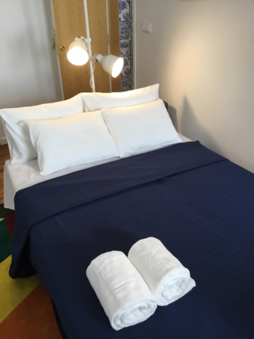 Quarto moderno em apartamento lisboeta com almofadas brancas, manta azul-marinho, toalhas enroladas e luminárias pendentes acima da cama