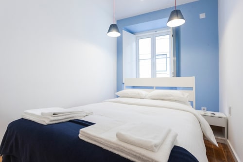 Quarto moderno em apartamento lisboeta com parede de destaque azul, roupa de cama branca, luminárias pendentes e janela clara com vista para
