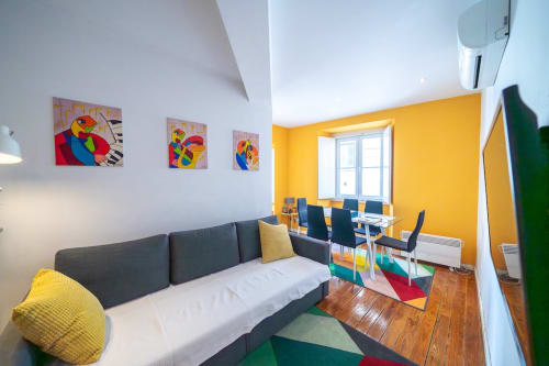 Apartamento moderno em Lisboa com obra de arte de papagaio colorido, sofá cinzento, paredes com acentos amarelos e zona de refeições com janela
