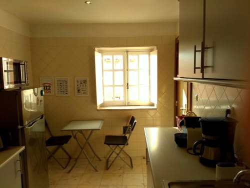 Cozinha clara e aconchegante no apartamento Bairro Alto, Lisboa, com mesa dobrável, cadeiras, janela, frigorífico e bancada em aço inoxidável