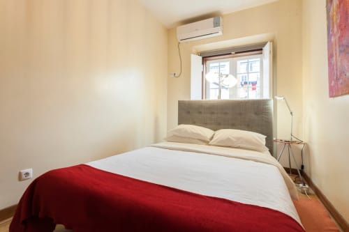 Quarto aconchegante no Heart Pulse Stays em Lisboa com cabeceira cinza, roupa de cama branca, manta vermelha, ar-condicionado e janela com vista da cidade