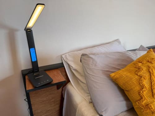 Luminária moderna com display digital azul sobre criado-mudo de madeira ao lado da cama na Villa Sereia Dourada.
