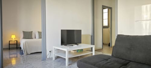Salón de apartamento en Eixample Barcelona con televisor, sofá gris y vista al dormitorio a través de mamparas de cristal