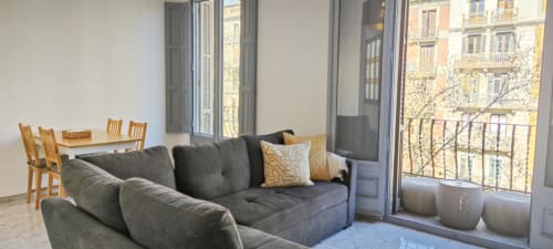 Moderno salón de apartamento en Barcelona con sofá gris, mesa de comedor de madera y vistas a la ciudad desde el balcón en el distrito de Eixample
