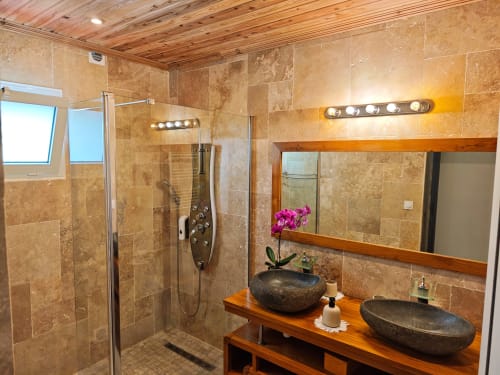 Salle de bain luxueuse du gîte La Kaz 57 à la Réunion avec carrelage en travertin, douche à effet pluie, doubles vasques et meuble de salle de bain en bois