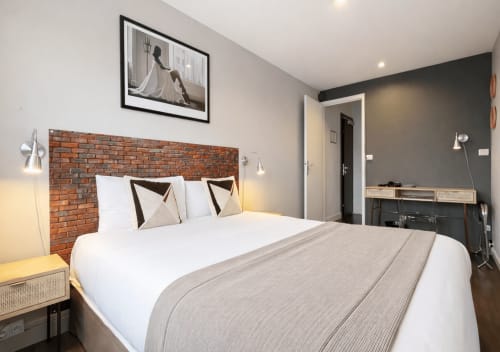 Chambre moderne de l'Hôtel Des Cèdres à Orléans avec tête de lit en brique, literie blanche, mur d'accent gris et vue sur la ville