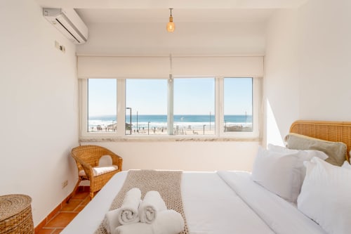 Bright beachfront bedroom in Aroeira, Portugal with sea view, white bedding, rattan furniture, and modern amenities