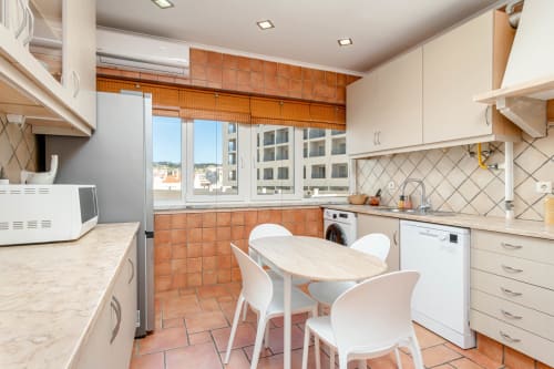 Modern apartment kitchen with dining area, orange tile backsplash, white cabinetry, sea view window in Charneca da Caparica