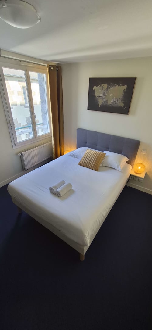 Chambre double moderne à l'hôtel Aux Genêts d'Or à Trévé avec lit blanc, tête de lit grise, mappemonde et vue sur la ville