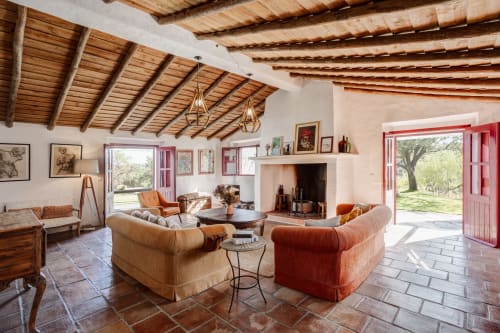 Sala de estar de villa alentejana rústica com vigas de madeira, lareira, soalho em terracota e portas vermelhas abertas para os jardins