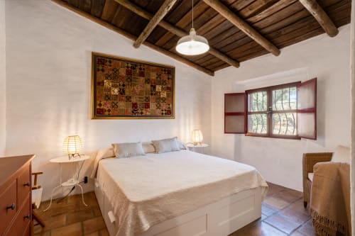 Quarto luminoso na villa Vale Clara, Alandroal, com teto de vigas aparentes, roupa de cama branca, cadeira de vime e tapeçaria tradicional