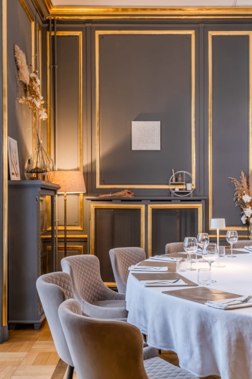 Élégante salle à manger de l'hôtel Ô Château à Hayange avec murs aux cadres dorés, nappes blanches et chaises matelassées beige.