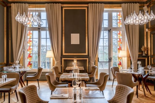 Élégante salle à manger de l'hôtel Ô Château à Hayange avec lustres modernes, grandes fenêtres, rideaux beige et design contemporain