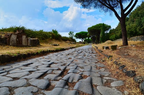 Antica strada romana in pietra con rovine presso le Stanze della Basilica a San Giovanni, Italia, circondata da cipressi in un paesaggio arido