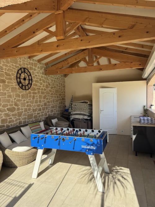 Spacieuse salle de loisirs du gîte avec baby-foot bleu, poutres en bois apparentes, murs en pierre et coin salon confortable
