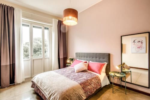 Camera da letto luminosa in appartamento verde Borghese con letto matrimoniale, accenti rosa, lampada a sospensione, balcone con vista sul giardino e design moderno