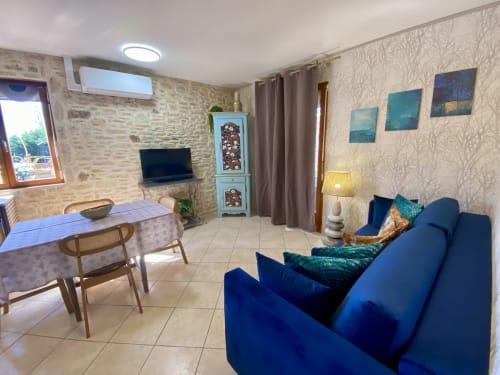 Séjour ouvert avec canapé bleu, salle à manger, télévision, murs en pierre et vue sur la piscine au gîte Le Petit Prince à Sarlat