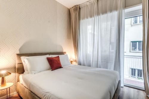 Camera da letto moderna con letto matrimoniale, biancheria bianca, cuscino rosso accento, carta da parati geometrica, porte balconate e luce naturale