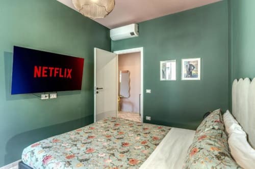 Camera da letto moderna con pareti verde salvia, biancheria floreale, TV a parete con Netflix, e porta aperta al bagno privato.