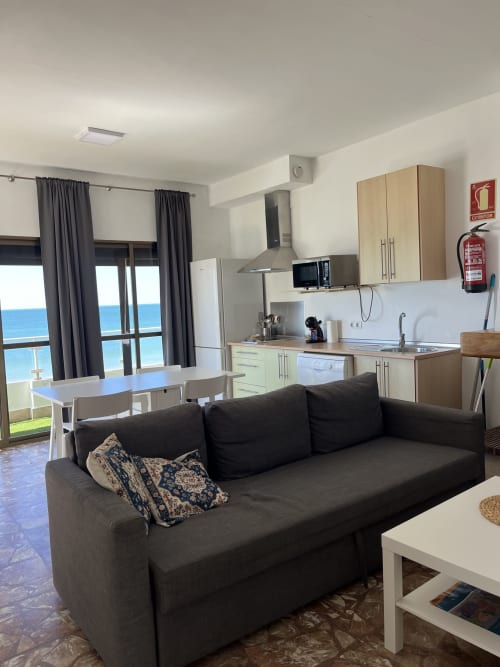Moderno salón con sofá negro, vistas al mar a través de puertas de terraza, cocina americana y decoración costera