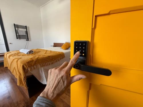 Mão inserindo código no painel da porta amarela vibrante da suíte Villa Sereia Dourada com cama e sistema de fechadura moderno